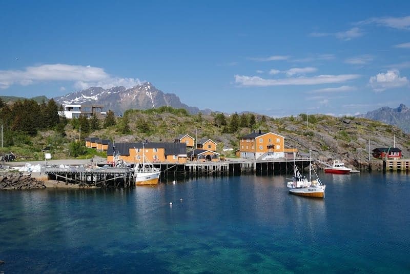 A serene coastal village with vibrant orange houses nestled by the water, surrounded by rugged mountains under a clear blue sky, ideal for travelers seeking a tranquil retreat in Norway's picturesque Lofoten Islands.
