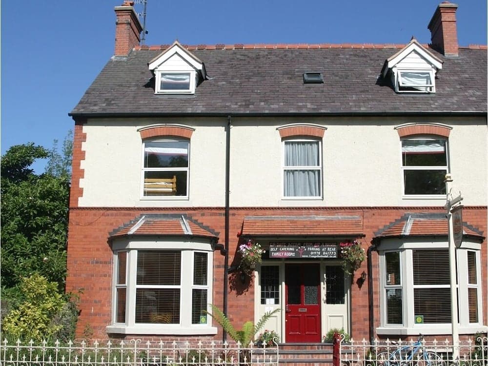A charming two-story Victorian-style guesthouse with red brick accents, bay windows, and a welcoming red door stands under a clear blue sky, ideal for travelers seeking cozy accommodations in a quaint setting.