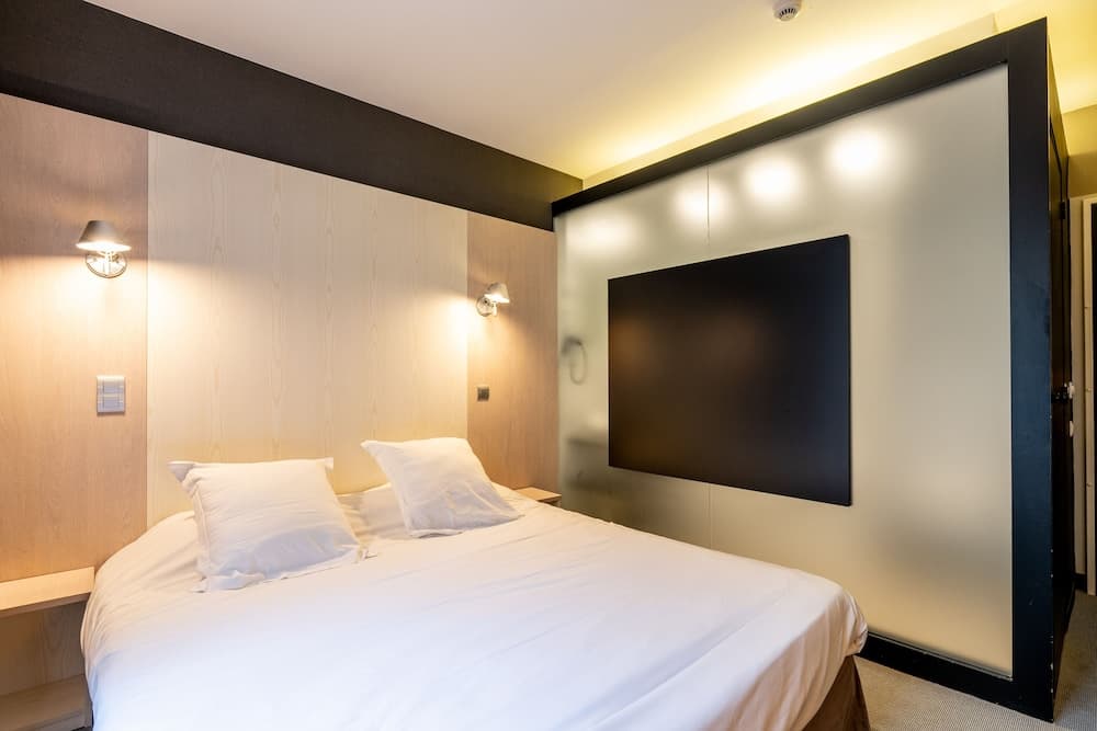 A cozy hotel room features a neatly made bed with white linens, wooden paneling, wall-mounted lamps, and a flat-screen TV, ideal for travelers seeking comfort and modern amenities.