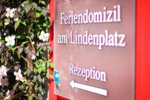A welcoming sign for "Feriendomizil am Lindenplatz" with an arrow pointing to the reception, set against a backdrop of blooming flowers, evokes a serene and inviting atmosphere ideal for travelers seeking a cozy accommodation experience.