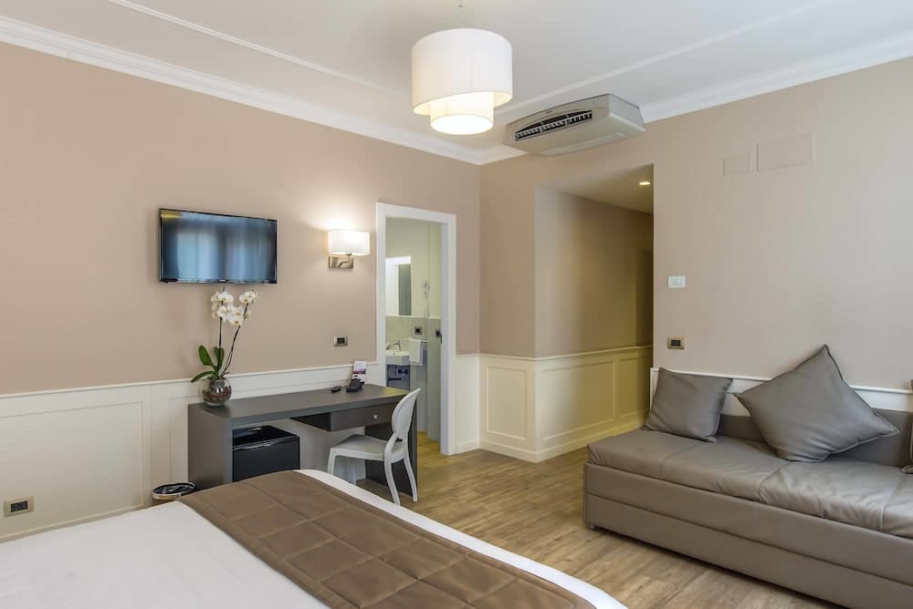A cozy hotel room features a neatly made bed, a comfortable sofa, a modern desk with a TV above it, and soft lighting, creating a welcoming atmosphere ideal for travelers seeking relaxation and convenience.