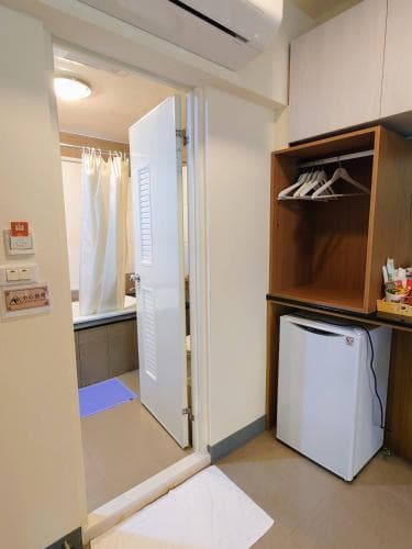 A cozy hotel room features a compact layout with a visible bathroom, wooden closet with hangers, mini-fridge, and soft lighting, ideal for travelers seeking comfort in a small space.