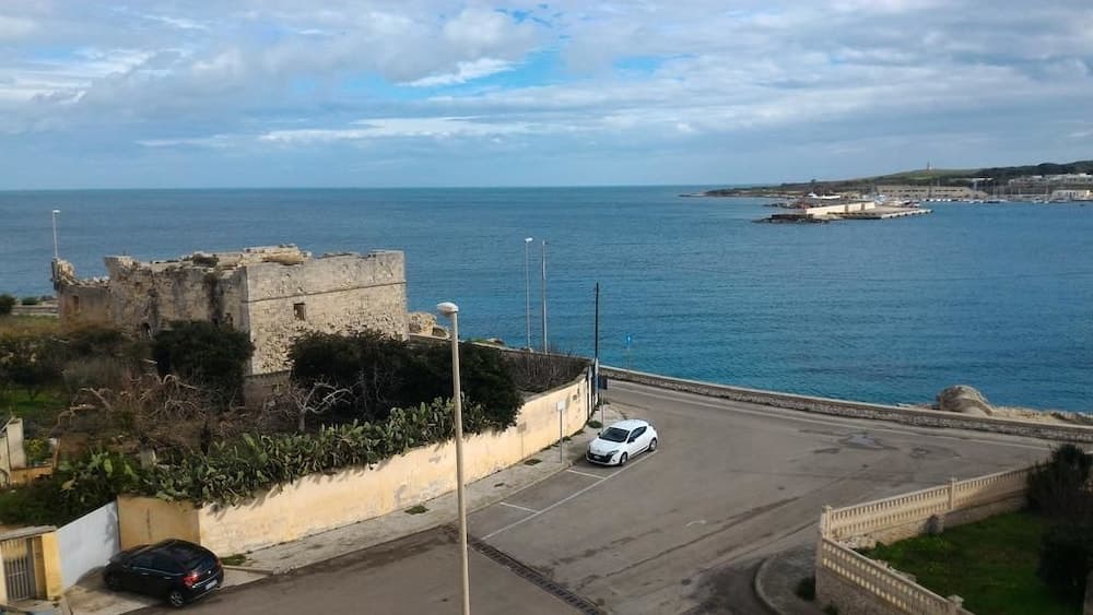 A serene coastal road lined with historic ruins and lush greenery offers travelers a picturesque route beside the calm blue sea, ideal for scenic drives and exploring cultural landmarks.