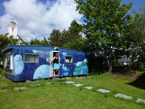 A vibrant blue caravan adorned with bubble art sits invitingly in a lush green garden, offering a quirky and cozy accommodation option for travelers seeking unique lodging experiences.