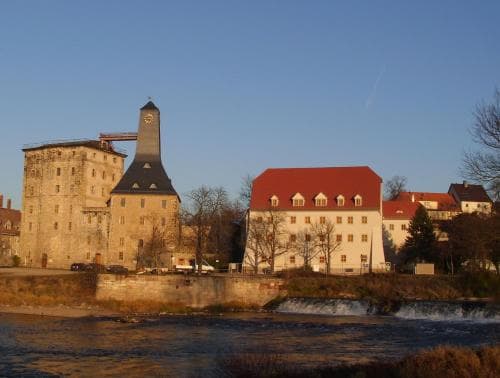 A serene riverside town features historic buildings with a prominent clock tower, cascading waterfalls, and warm evening light, ideal for cultural exploration and tranquil getaways.