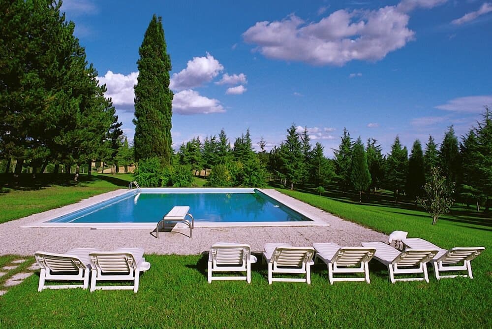 A serene outdoor pool surrounded by lush greenery and tall trees under a bright blue sky, featuring white lounge chairs on a grassy lawn, ideal for a relaxing vacation getaway.