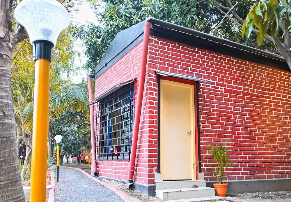 A charming red brick building with a yellow door and window grills sits amidst lush greenery, creating a serene park atmosphere ideal for leisurely strolls and family outings.