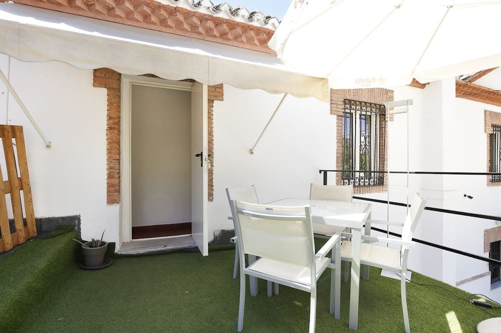 A cozy outdoor patio with white furniture under an umbrella, artificial turf flooring, and a brick-accented wall creates a serene retreat ideal for vacation rentals or leisurely stays.