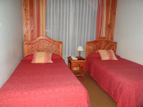 A cozy hotel room features two single beds with red bedding, wooden headboards, and a small bedside table with a lamp, ideal for travelers seeking a comfortable and budget-friendly stay.