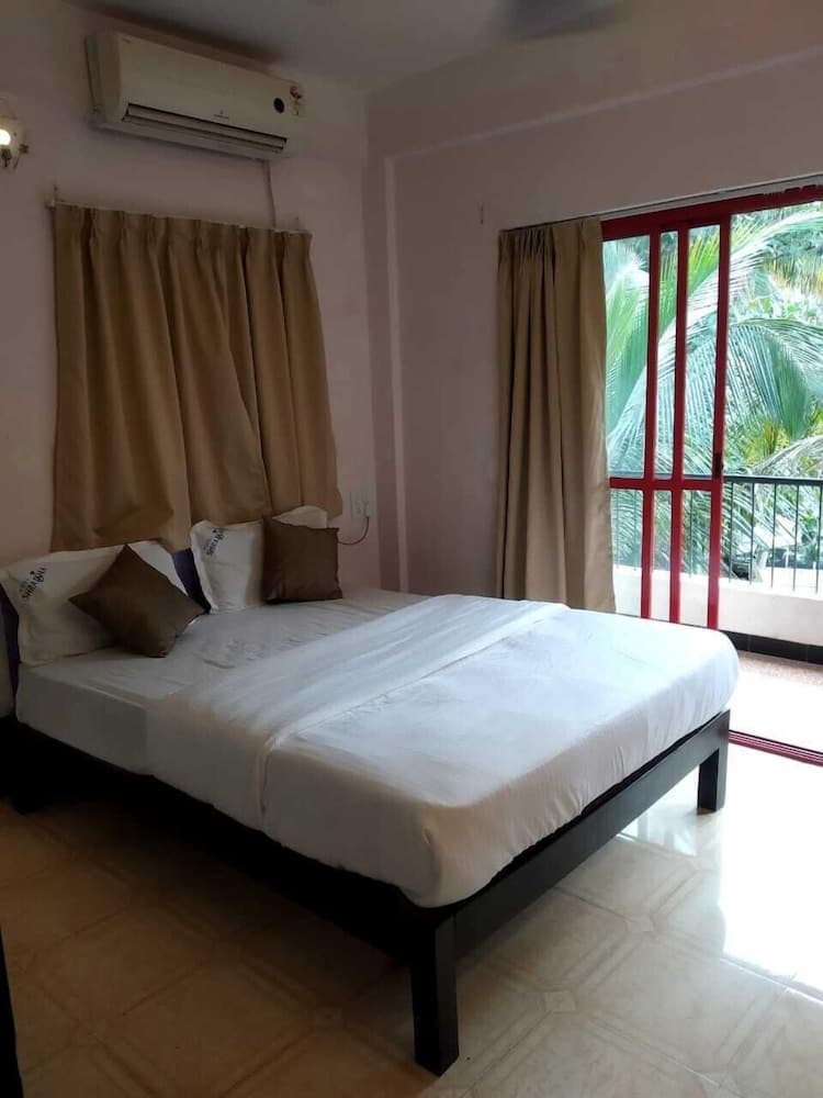 A cozy hotel room with a neatly made bed, beige curtains, and a red-framed balcony door offering a view of lush palm trees, ideal for travelers seeking relaxation in a tropical setting.