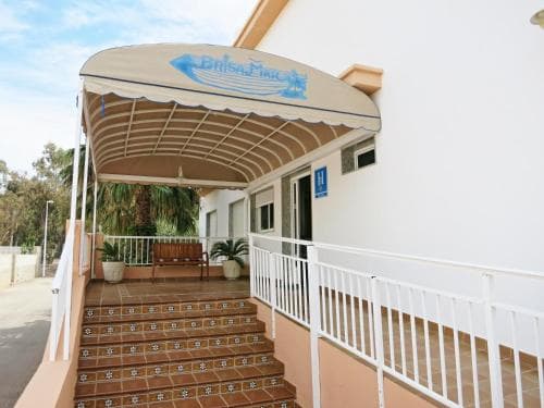 A sunny Mediterranean hotel entrance features tiled stairs, a welcoming awning, and lush greenery, ideal for travelers seeking a cozy and relaxed stay.