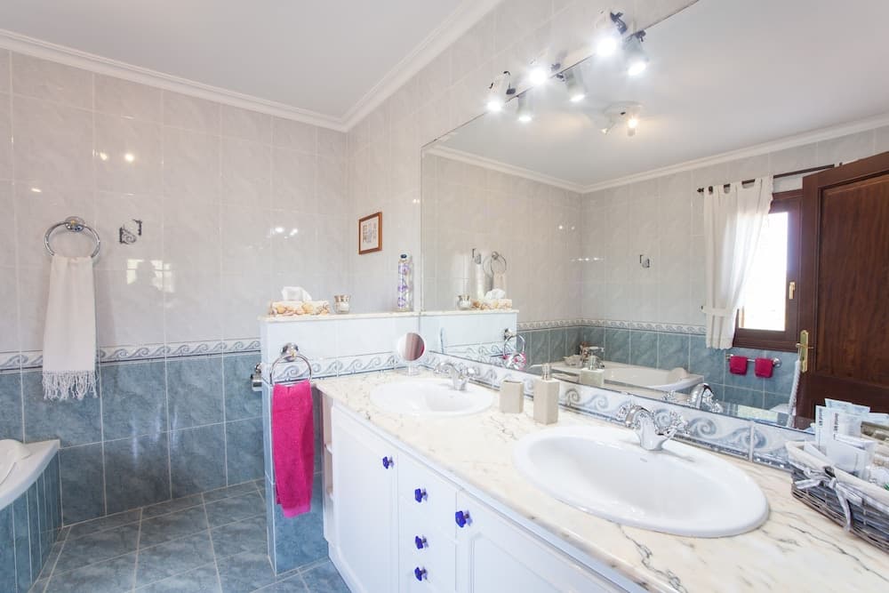 A serene and spacious bathroom features dual sinks, marble countertops, blue-tiled walls, and a large mirror reflecting natural light from a window, ideal for travelers seeking comfort and luxury in their accommodations.