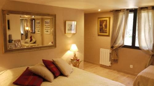 A cozy hotel room features a neatly made bed with red accents, a large mirror reflecting the space, soft lighting from a bedside lamp, and a window with curtains, ideal for travelers seeking comfort and relaxation.