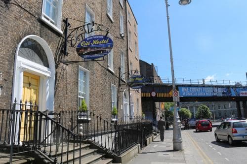 A charming urban street scene features a quaint brick building with a "Backpackers City Hotel" sign, evoking a welcoming atmosphere for travelers seeking budget-friendly accommodations in a lively city setting.