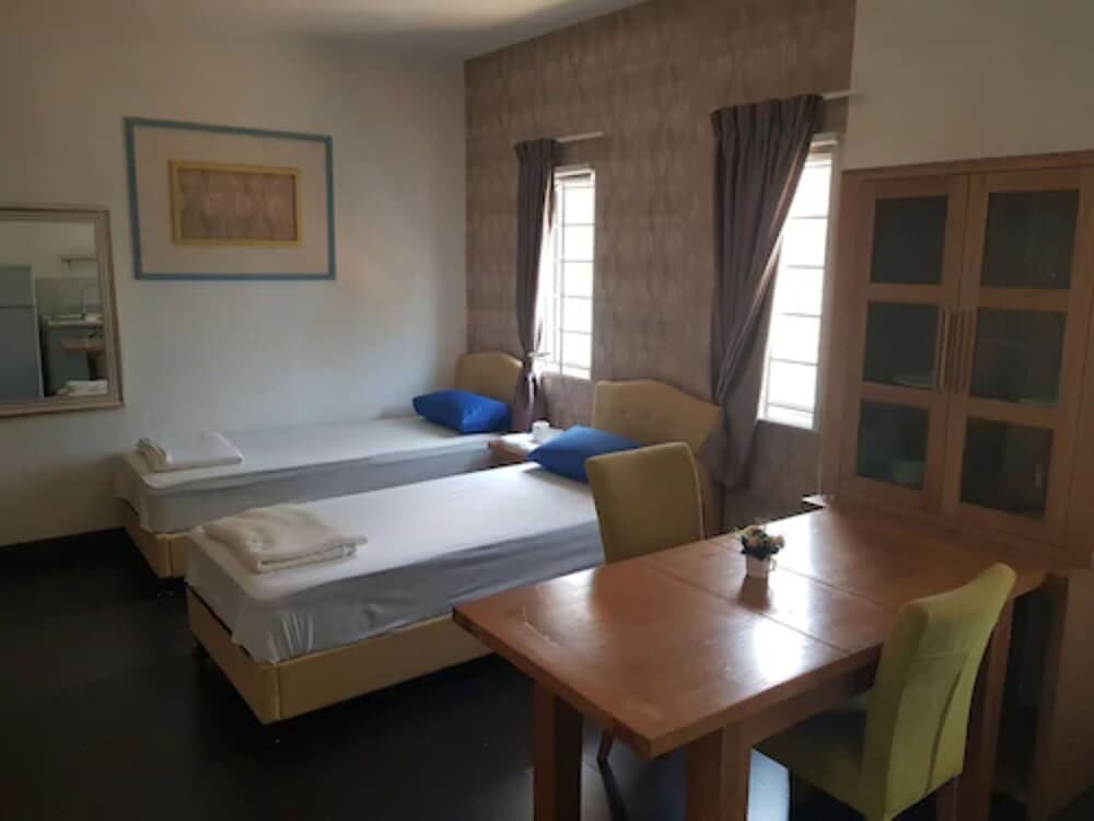 A cozy studio apartment features two single beds, a wooden dining set, and a glass-front cabinet, creating a welcoming atmosphere ideal for budget travelers seeking comfort and functionality.