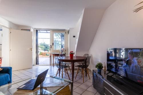 A cozy attic apartment features a modern living space with a glass coffee table, laptop, flat-screen TV, and a bright balcony view, ideal for travelers seeking comfort and relaxation in a stylish urban retreat.