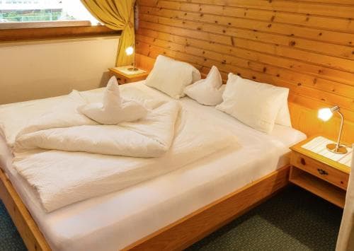 A cozy wooden cabin bedroom with neatly arranged white bedding, soft lighting, and a bedside table creates a warm and inviting atmosphere ideal for travelers seeking relaxation and comfort.