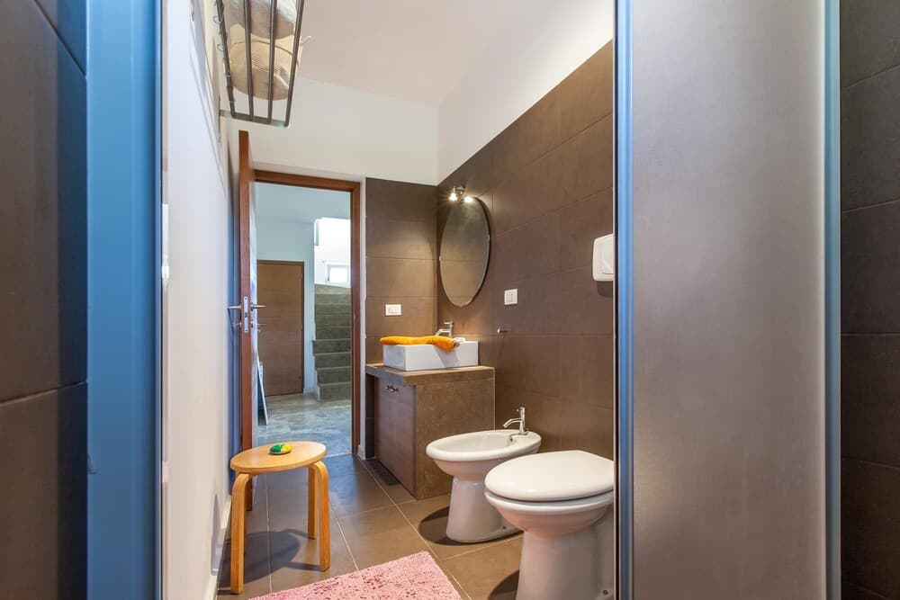 A modern bathroom with brown tiled walls, a sleek sink, bidet, toilet, and wooden stool creates a cozy and functional space ideal for travelers seeking comfort in accommodations.
