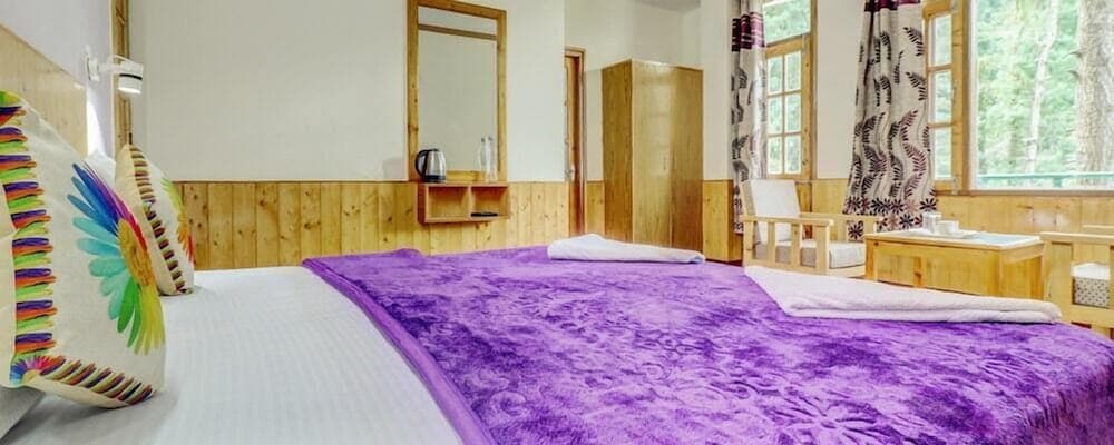 A cozy cabin bedroom with wooden walls, a vibrant purple bedspread, colorful pillows, and large windows overlooking lush greenery, ideal for travelers seeking a serene retreat in nature.