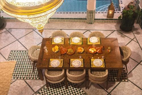 A cozy indoor dining area features a wooden table set with colorful plates and traditional Moroccan decor, including tiled floors and ornate lanterns, ideal for travelers seeking an authentic cultural culinary experience.