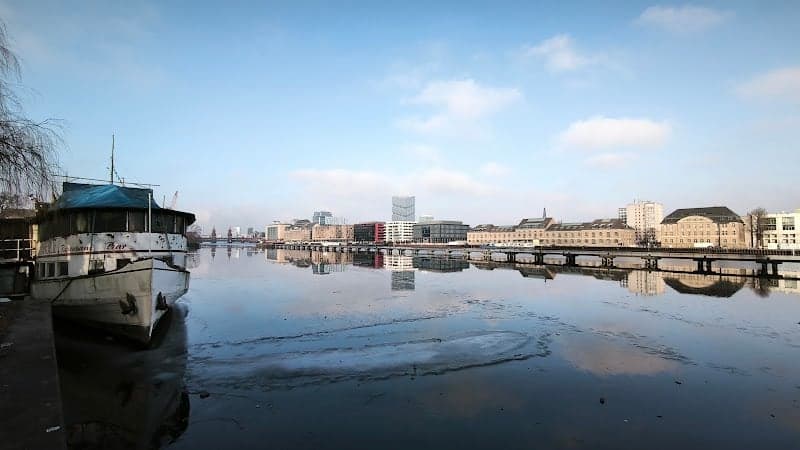 A serene winter scene captures the icy Spree River in Berlin's Friedrichshain-Kreuzberg district, featuring the historic MS Hoppetosse ship and urban skyline reflections, ideal for a tranquil city break adventure.