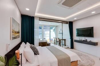 A cozy modern bedroom with plush bedding, teal curtains, and a flat-screen TV overlooks a bright patio area, ideal for travelers seeking comfort and relaxation in a stylish urban retreat.