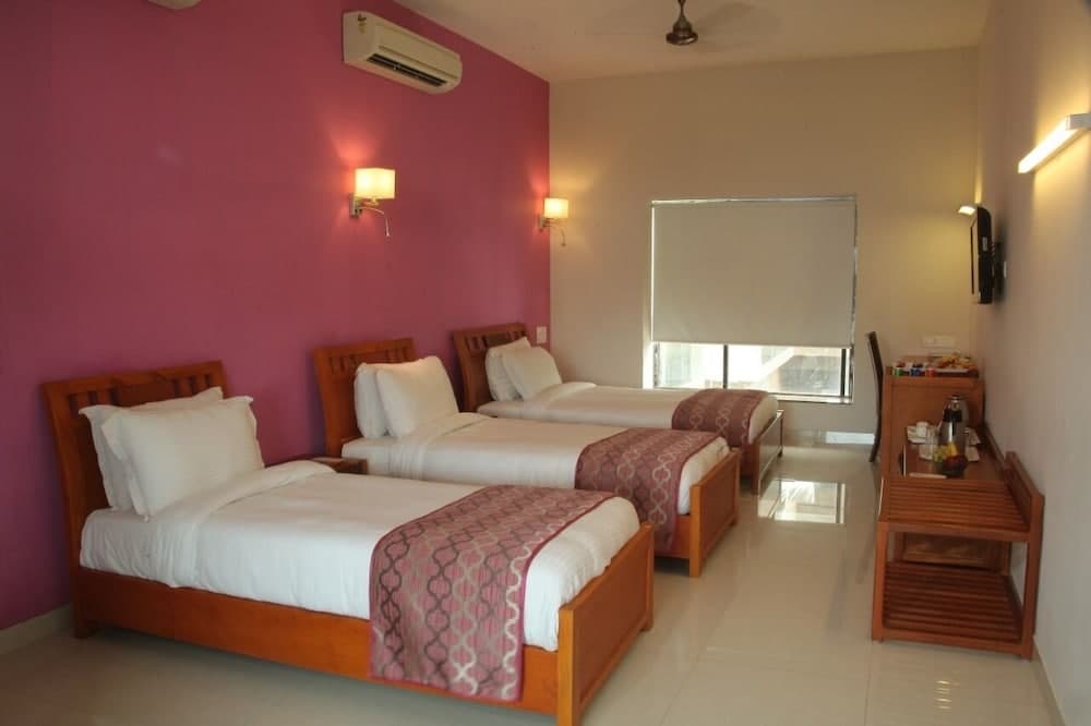 A cozy hotel room with three neatly arranged beds, a vibrant pink accent wall, wooden furnishings, and natural light streaming through a large window, ideal for travelers seeking comfort and simplicity.