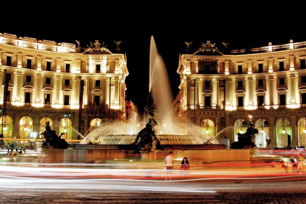 A vibrant night scene captures a grand fountain illuminated by historic buildings, evoking a romantic European square atmosphere ideal for evening strolls and photography enthusiasts.