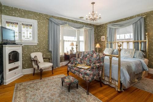 A cozy, vintage-inspired bedroom with ornate wallpaper, a brass bed, floral armchair, and stained glass window exudes charm and comfort, ideal for travelers seeking a quaint and elegant accommodation experience.