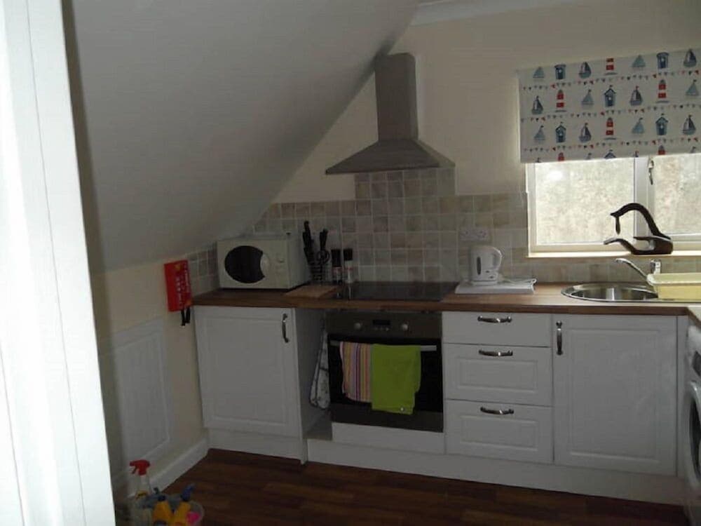 A cozy attic kitchen with white cabinets, tiled backsplash, and nautical-themed window decor offers a charming space for travelers seeking self-catering accommodations.