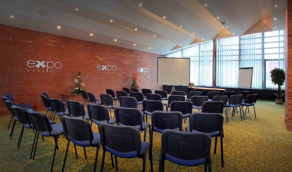 A modern conference room with rows of chairs facing a projector screen, set against a brick wall with "expo hotel" branding, ideal for business travelers seeking professional meeting spaces.