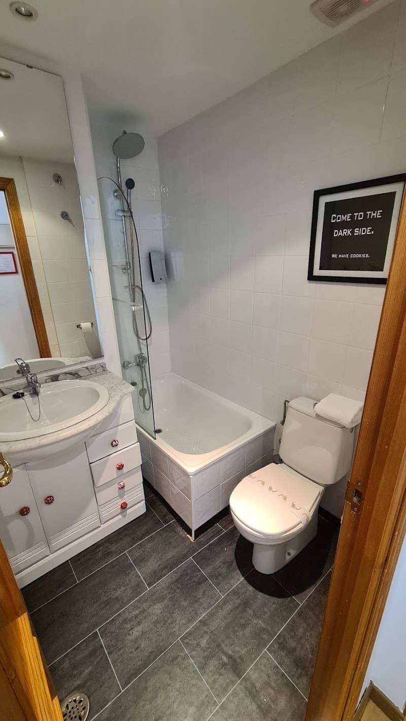 A cozy bathroom inside Plaza Sierra Nevada features white tiled walls, a bathtub with a shower, a sink with red-knobbed drawers, and a humorous wall sign, ideal for travelers seeking comfort in Granada, Spain.