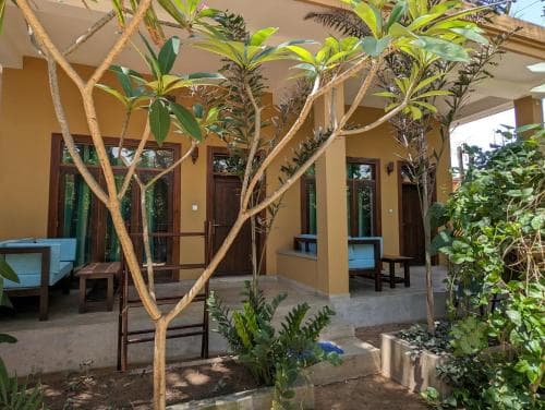 A serene tropical courtyard features lush greenery and wooden furniture outside a cozy, yellow-walled villa, ideal for travelers seeking a peaceful retreat.