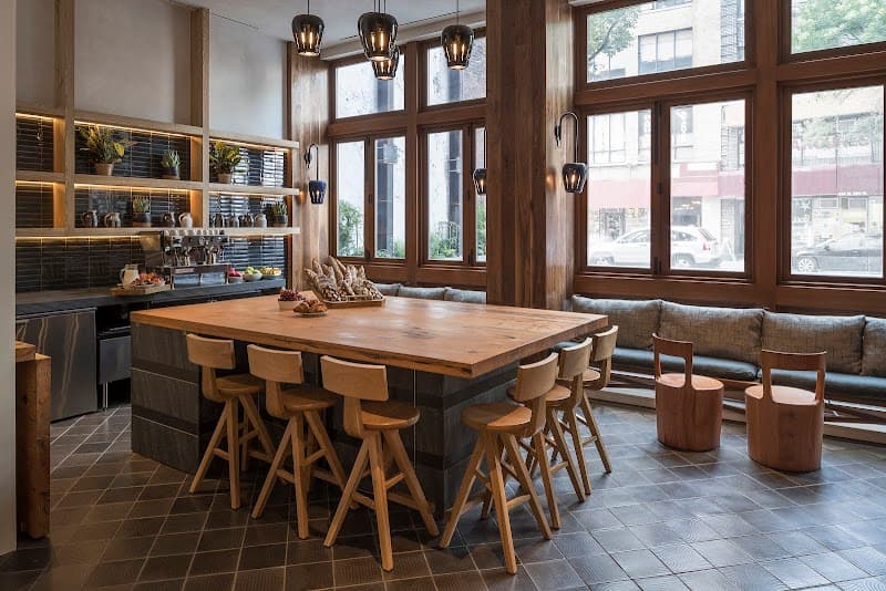A cozy urban retreat at the Made Hotel in New York features a rustic wooden communal table surrounded by stools, set against large windows with city views, ideal for travelers seeking a warm and inviting space to relax or work.