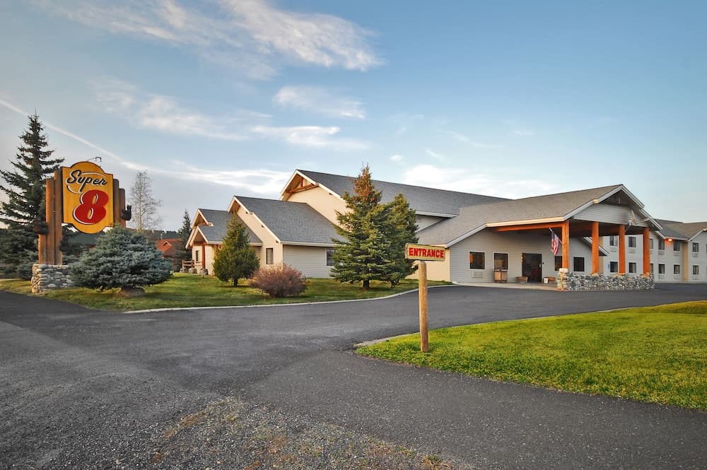 A welcoming Super 8 motel entrance features a well-maintained lawn, pine trees, and a clear blue sky, ideal for travelers seeking comfortable lodging in a serene suburban setting.