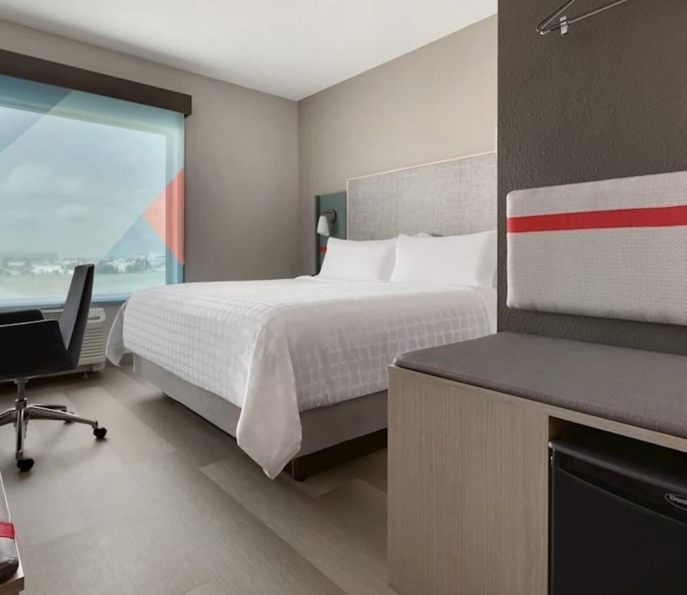 A modern hotel room with a cozy bed, sleek bench, and large window offering an airport view, ideal for travelers seeking comfort and convenience during layovers or short stays.
