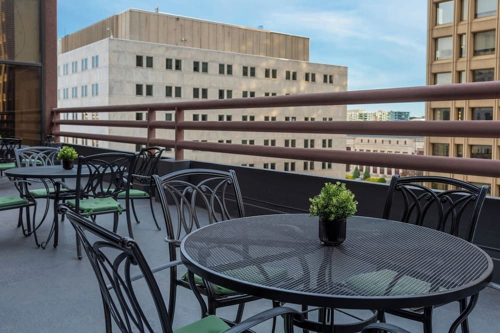 A serene rooftop patio features black metal tables adorned with small green plants, surrounded by urban buildings under a clear sky, ideal for city travelers seeking a peaceful dining or relaxation spot.