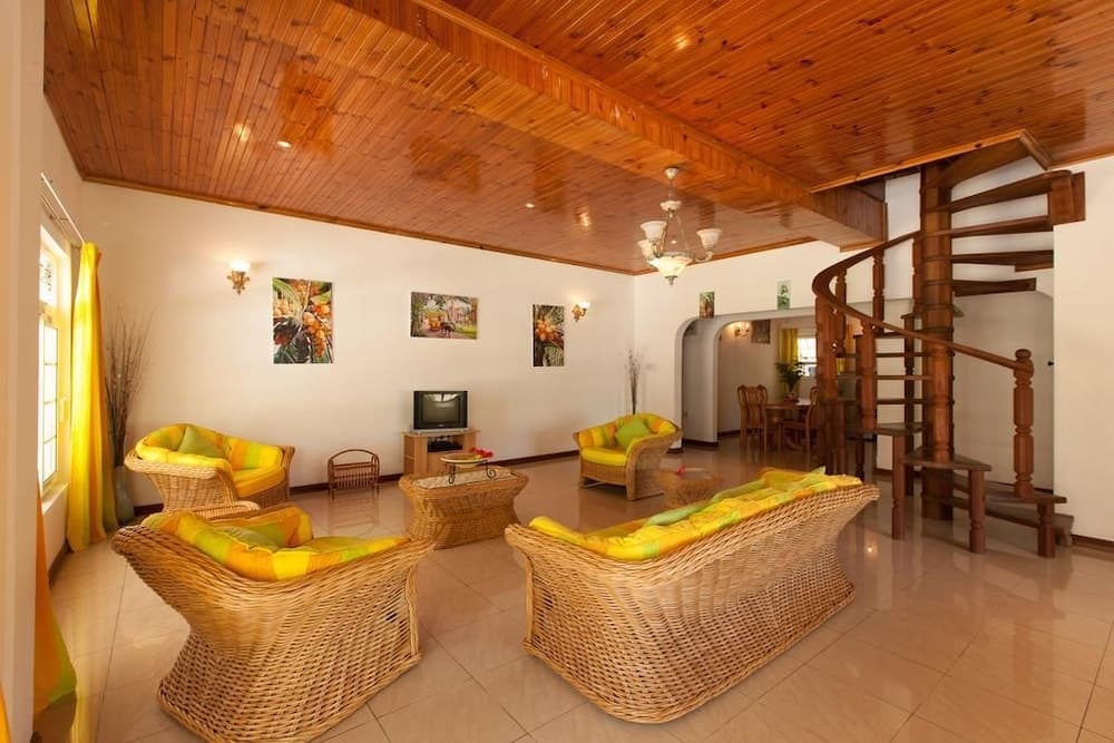 A cozy living room with wicker furniture, vibrant yellow cushions, wooden ceilings, and a spiral staircase exudes warmth and comfort, ideal for travelers seeking a relaxing tropical retreat.