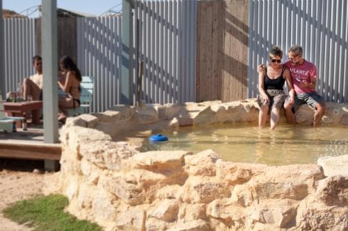 A couple relaxes in a natural stone hot spring at an outdoor spa, enjoying the serene atmosphere with lounge chairs and wooden structures nearby, perfect for a tranquil getaway.