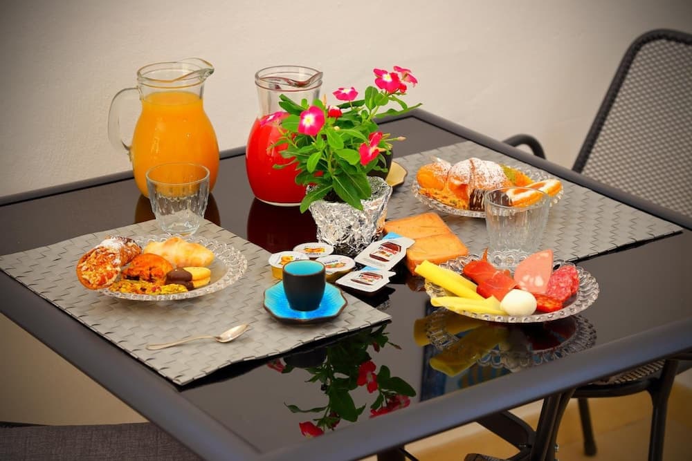 A cozy outdoor dining setup features a vibrant breakfast spread with pastries, fruits, cheese, and orange juice on a reflective table, complemented by a blooming flower pot, ideal for travelers seeking a charming morning experience.