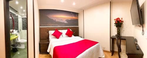 A cozy hotel room features a neatly made bed with red accents, a scenic wall mural, and an adjacent bathroom, creating a welcoming atmosphere ideal for travelers seeking comfort and convenience.