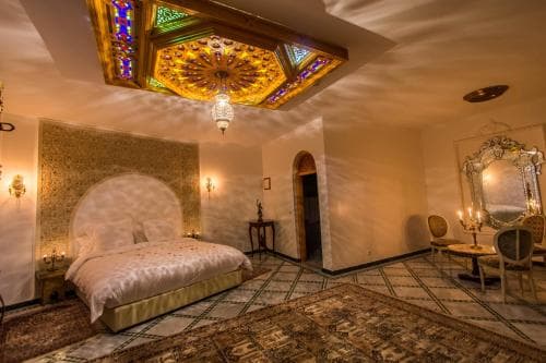A luxurious bedroom with ornate Moroccan-style decor, featuring a glowing stained-glass ceiling light, plush bed, intricate rugs, and elegant furnishings, creates an inviting atmosphere ideal for a romantic getaway or cultural immersion experience.