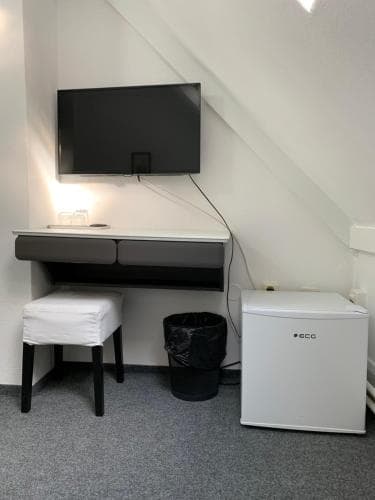 A cozy attic room features a sleek desk setup with a mounted TV, a mini fridge, and a simple chair, ideal for travelers seeking a functional and minimalist workspace in a compact space.
