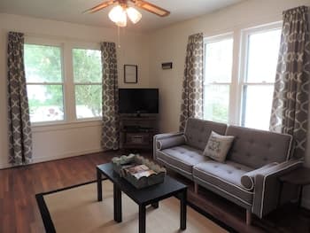 A cozy living room with hardwood floors, large windows draped in patterned curtains, a comfortable sofa, and a coffee table creates an inviting atmosphere ideal for a relaxing getaway or home staging.