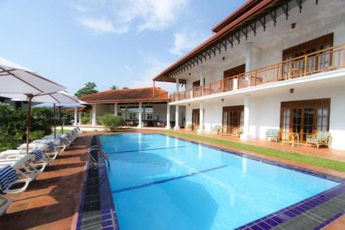 A serene resort scene features a sparkling pool lined with lounge chairs under umbrellas, set against a backdrop of a two-story building with balconies, ideal for a relaxing vacation getaway.