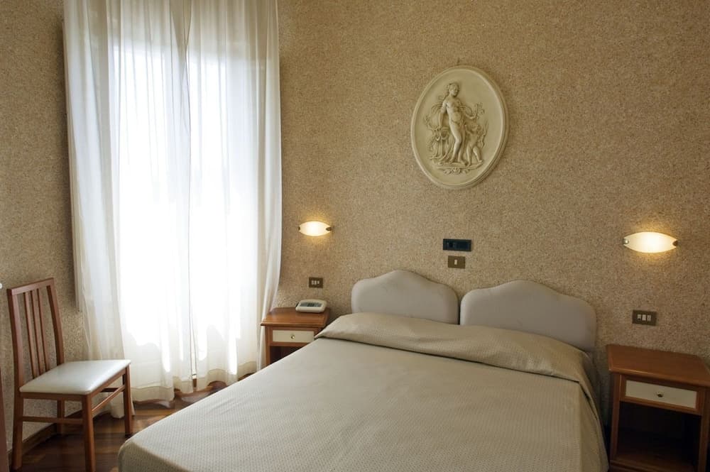 A cozy hotel room features a neatly made bed flanked by wooden nightstands, soft lighting, a serene wall sculpture, and sheer curtains diffusing natural light, ideal for travelers seeking comfort and tranquility.