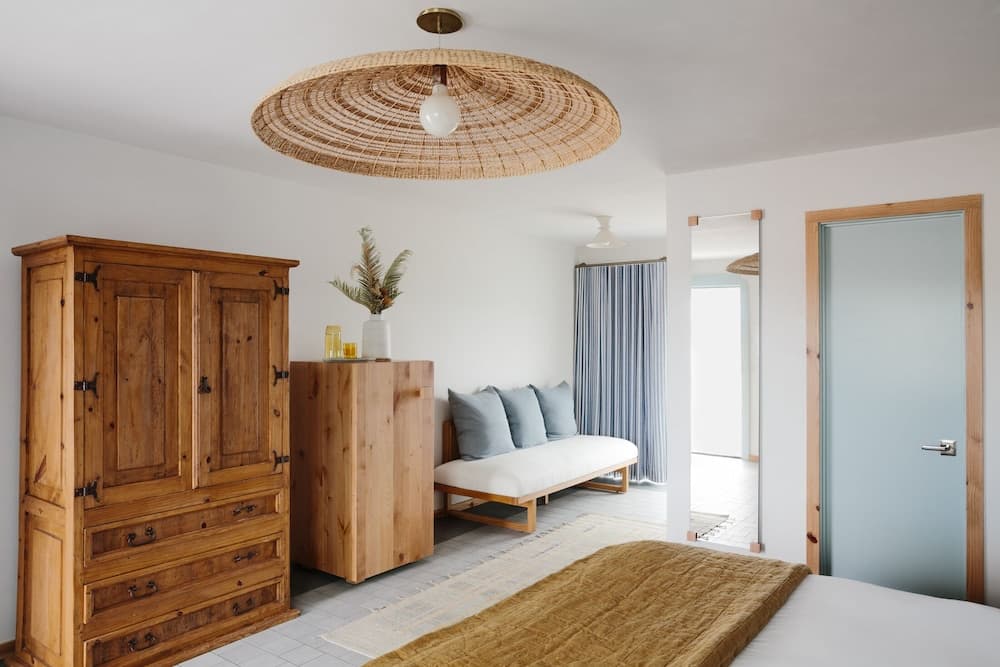 A serene bedroom with white walls, wooden furniture, a woven ceiling light, and a cozy bed setup evokes a tranquil retreat ideal for relaxation and leisure stays.