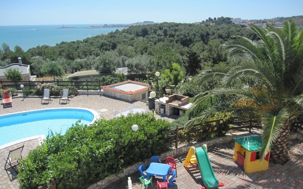 A serene coastal villa features a vibrant poolside area with lounge chairs, lush greenery, a children's play area, and panoramic sea views, ideal for family vacations seeking relaxation and recreation.