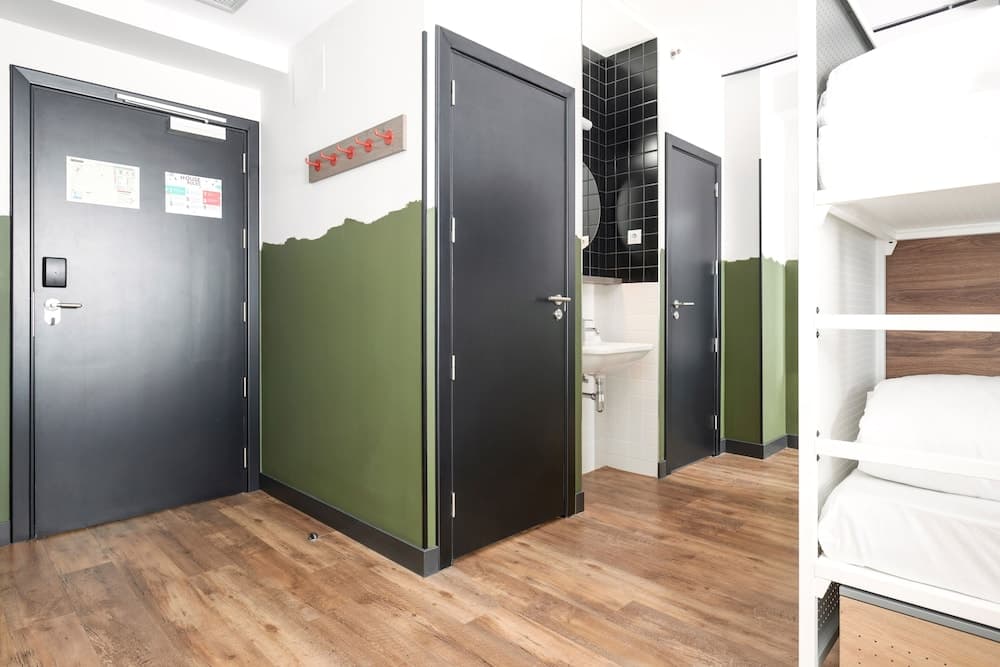 A cozy hostel room with bunk beds, wooden flooring, green-painted walls, and a bathroom in the background offers travelers a clean and modern accommodation option.