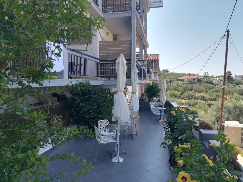 A serene outdoor terrace with white umbrellas, chairs, and lush greenery offers a tranquil spot for relaxation amidst a picturesque village setting, ideal for travelers seeking a peaceful retreat.